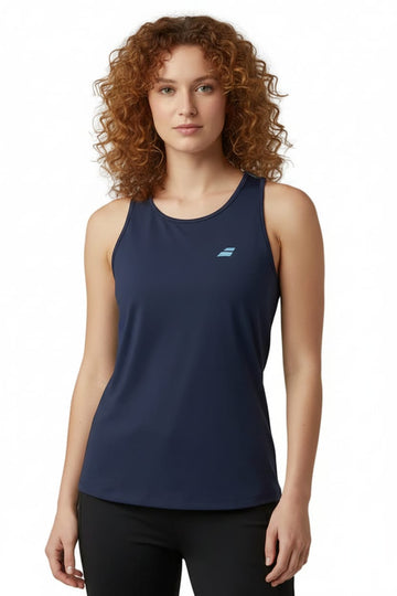 A woman with curly hair models the Babolat Drive Womens Tennis Tank Top - Blue—a sleeveless tennis top by Babolat in navy with a small blue logo on the left chest, made from lightweight Fiber Dry polyester—posing against a plain white background.