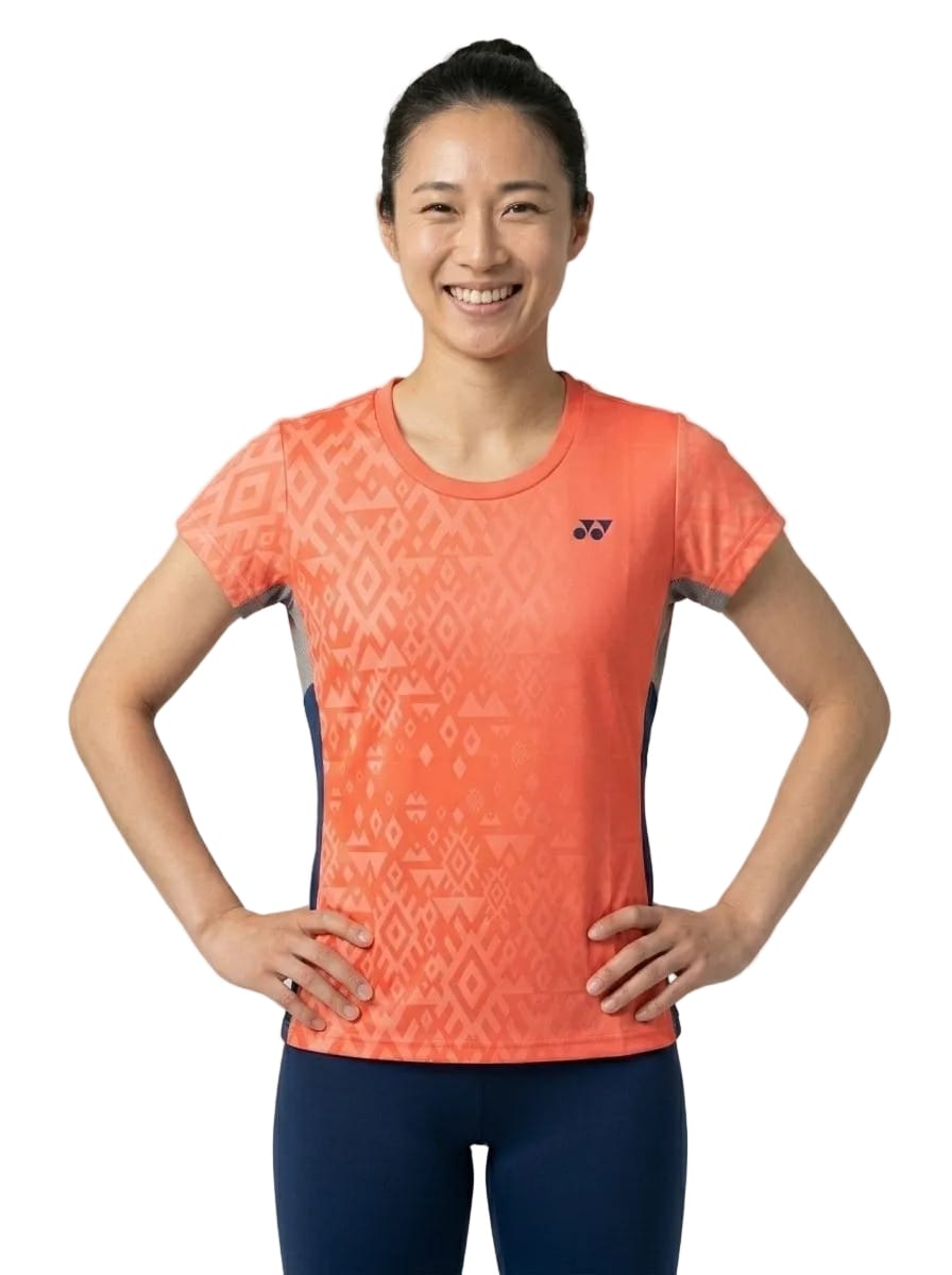 A smiling woman with dark hair in a ponytail is standing with her hands on her hips against a plain white background, wearing the Yonex YTL11 Women’s Tennis T-Shirt in Sunset and blue pants. The lightweight performance fabric of the Yonex top features an orange patterned design.