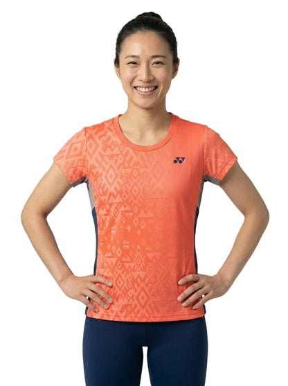 A smiling woman with dark hair in a ponytail is standing with her hands on her hips against a plain white background, wearing the Yonex YTL11 Women’s Tennis T-Shirt in Sunset and blue pants. The lightweight performance fabric of the Yonex top features an orange patterned design.