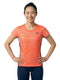 A smiling woman with dark hair in a ponytail is standing with her hands on her hips against a plain white background, wearing the Yonex YTL11 Women’s Tennis T-Shirt in Sunset and blue pants. The lightweight performance fabric of the Yonex top features an orange patterned design.