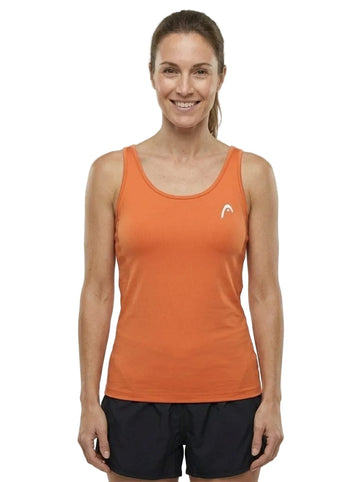 A woman stands smiling, wearing the HEAD Spirit II Womens Tennis Tank Top - CN by HEAD in orange—a sleeveless athletic top featuring cooling technology and a small white logo—paired with black shorts. She faces forward against a plain white background.