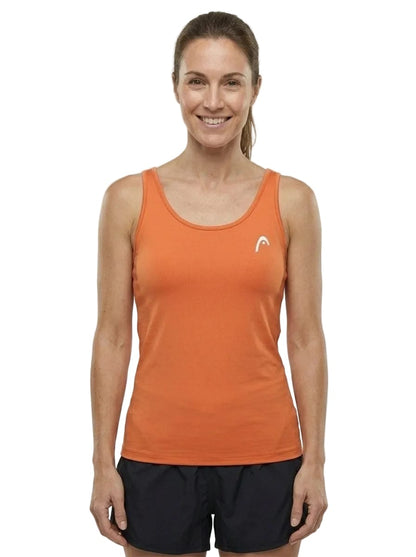 A woman stands smiling, wearing the HEAD Spirit II Womens Tennis Tank Top - CN by HEAD in orange—a sleeveless athletic top featuring cooling technology and a small white logo—paired with black shorts. She faces forward against a plain white background.