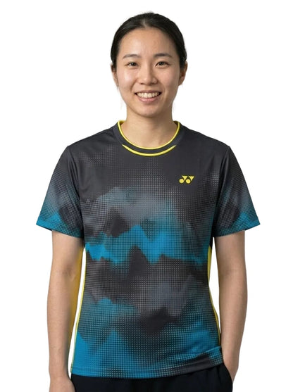 A person with long dark hair tied in a ponytail is wearing the Yonex YTL2025B Womens Tennis T-Shirt in black, featuring blue patterns and yellow accents on breathable sports fabric. She stands facing the camera and smiles against a plain white background.