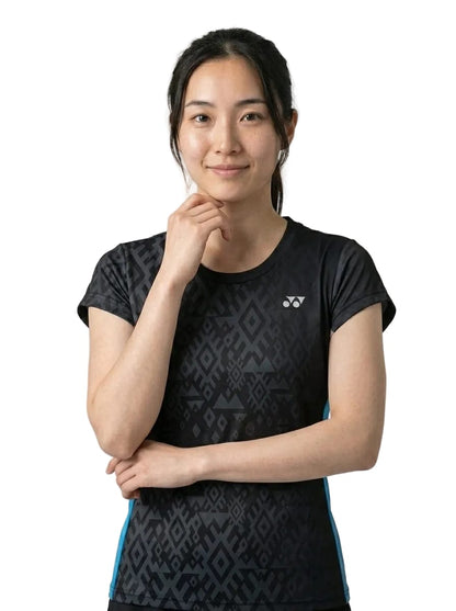A woman stands against a white background, wearing the Yonex YTL11 Womens Tennis T-Shirt in black. Crafted from lightweight performance fabric and featuring short sleeves, the t-shirt displays a subtle black pattern. She smiles softly, touching her chin thoughtfully with one hand.