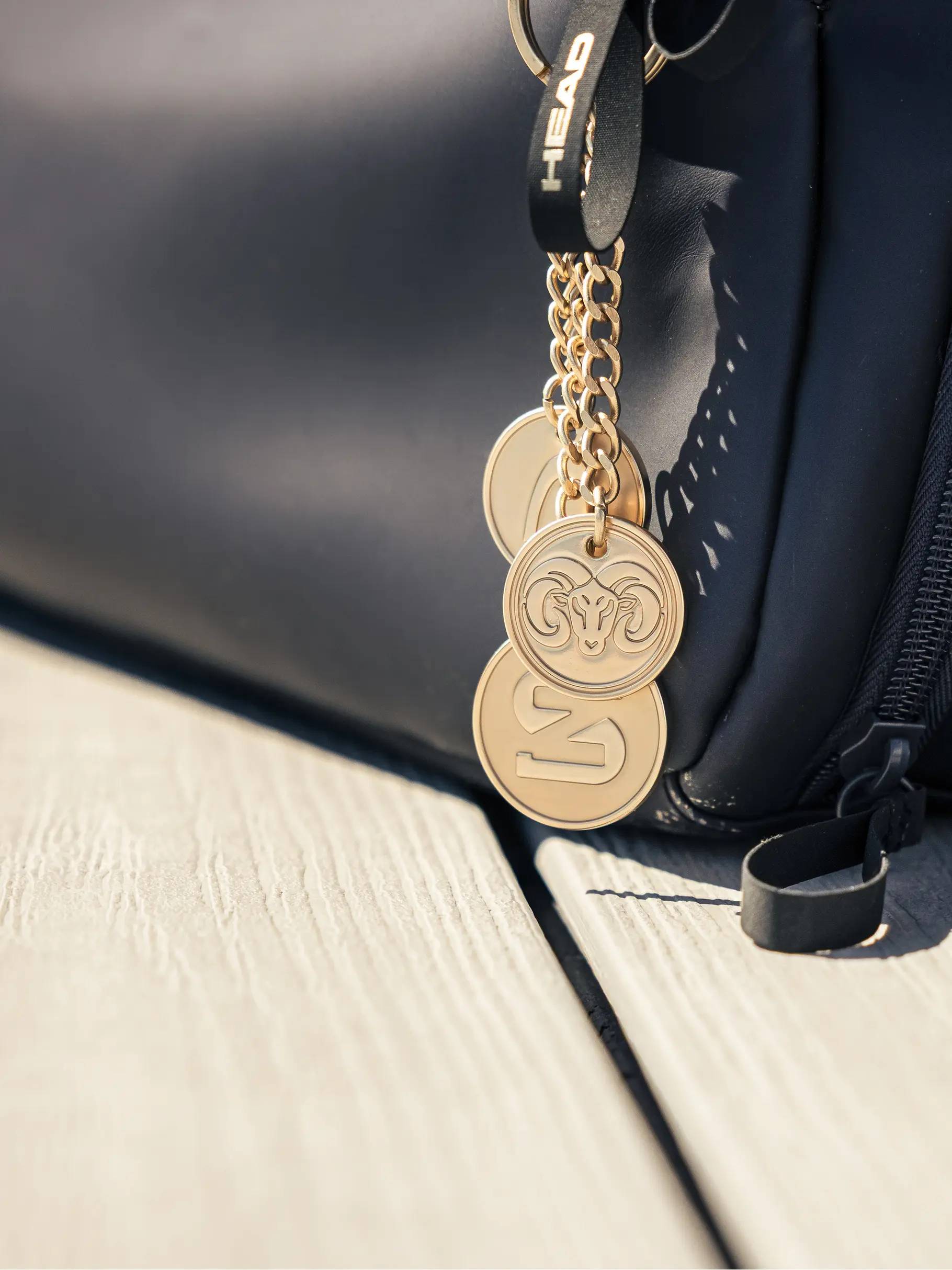 Close-up of gold keychains with engraved designs attached to a navy HEAD Pro X Zverev LTD Tennis Duffle Bag L, resting on a light wooden surface.