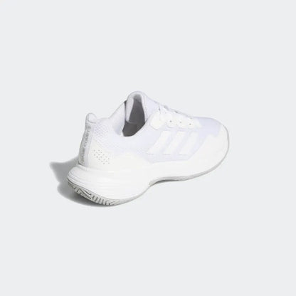 The adidas GameCourt 2 Women's Tennis Shoes in white are displayed at an upward angle, emphasizing their striped design and textured hard court outsole. The light gray background accentuates the sleek style of these shoes by adidas.