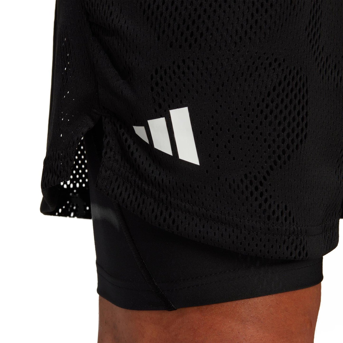 Close-up of a person wearing black adidas Melbourne 2in1 Men's 7 Inch Tennis Shorts, showcasing the iconic white, stylized three-stripe logo. The AEROREADY fabric offers enhanced ventilation with its perforated design against a plain white background.