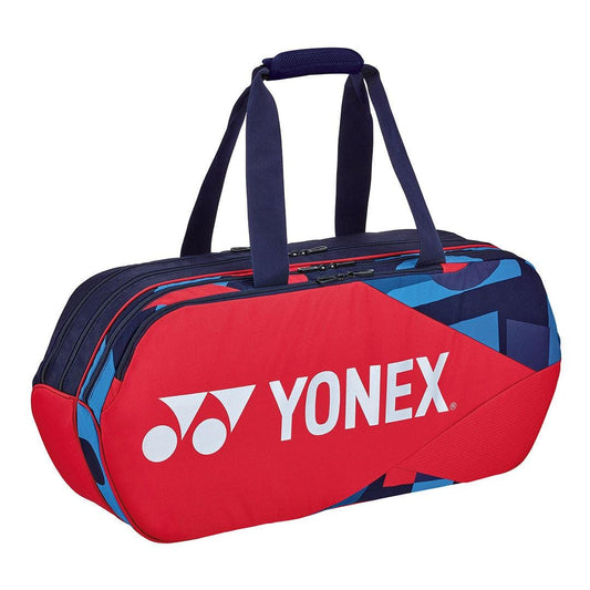 The Yonex 92231WEX Pro Tournament Bag - Scarlet showcases a vibrant geometric design in red and blue, prominently featuring the Yonex logo in white on the side. Designed to professional standards, it includes two durable navy handles with zippered compartments, complemented by accents of scarlet red.