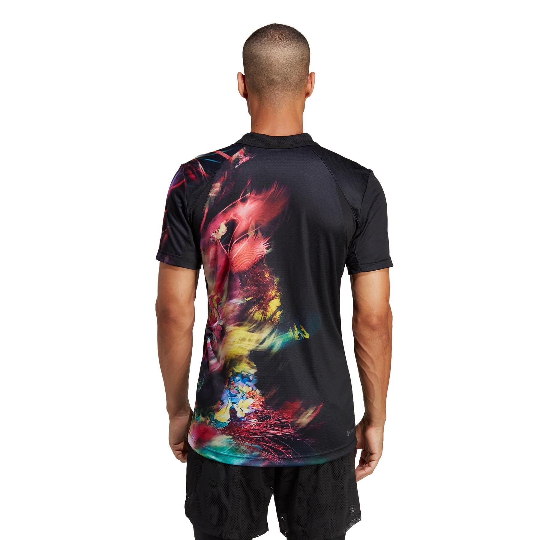 A person dons the adidas Melbourne Men's Freelift Tennis Polo Shirt in black with vibrant, abstract multicolored patterns visible from the back. They have short hair and complement their look with FreeLift black shorts. The scene is set against a plain white background.