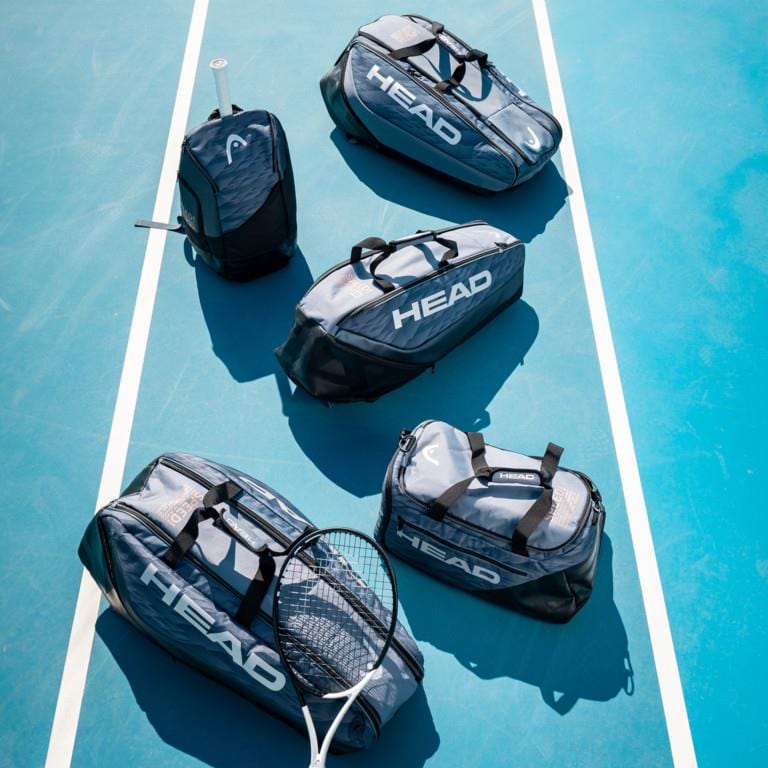 Five black and gray sports bags from HEAD are neatly arranged on a blue tennis court, showcasing a padded racket compartment. A tennis racket lies beside them, while the HEAD Djokovic Tennis Backpack - Black / Grey, featuring a ventilated shoe compartment, stands upright on the left. The scene is bright and well-lit.