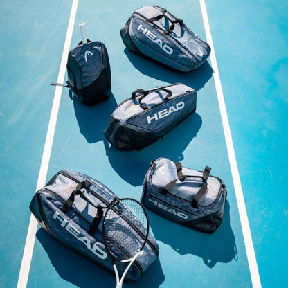 Five black and gray sports bags from HEAD are neatly arranged on a blue tennis court, showcasing a padded racket compartment. A tennis racket lies beside them, while the HEAD Djokovic Tennis Backpack - Black / Grey, featuring a ventilated shoe compartment, stands upright on the left. The scene is bright and well-lit.