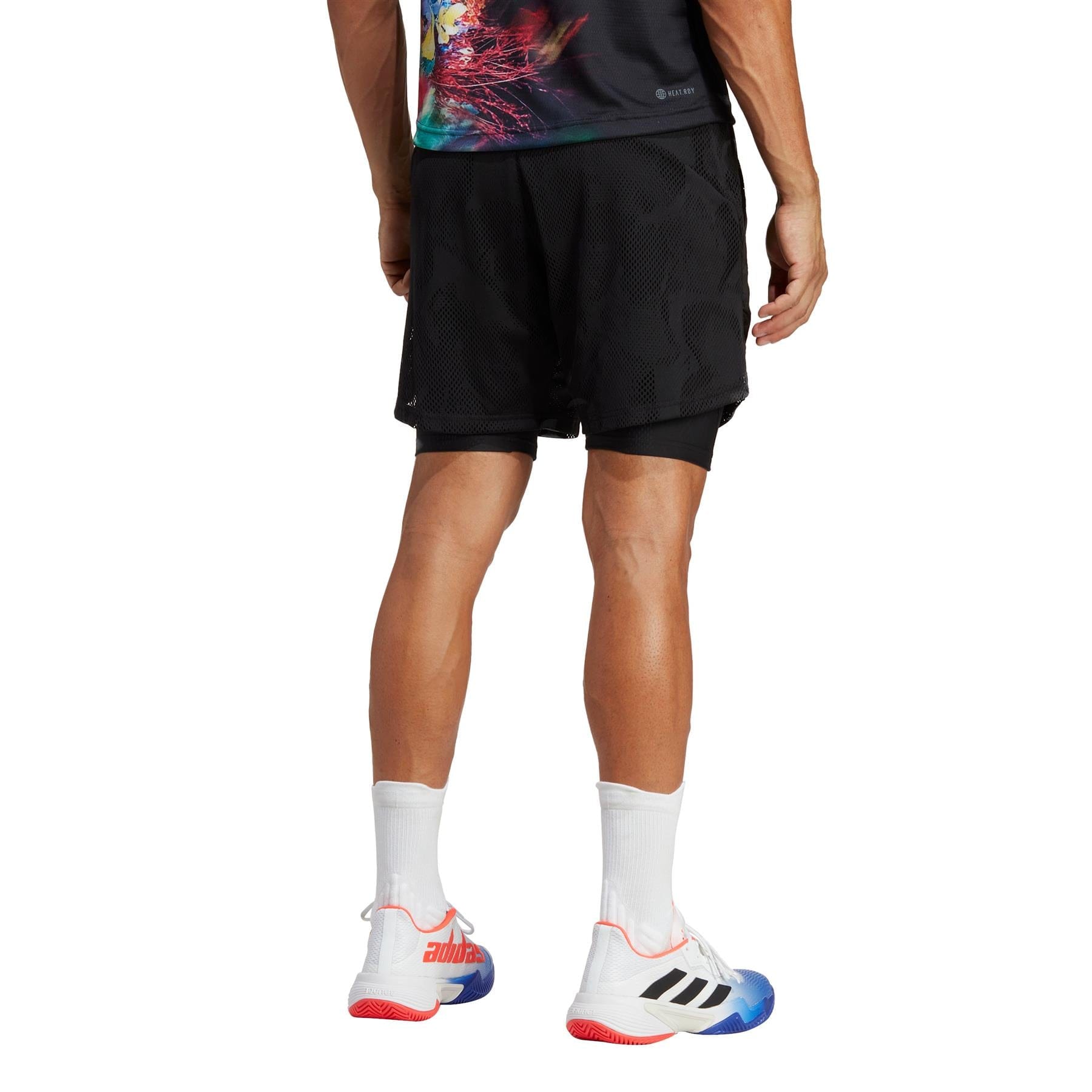 A person stands facing away, wearing a colorful T-shirt and adidas Melbourne 2in1 Men's 7 Inch Tennis Shorts in black, featuring AEROREADY technology. They are complemented by white athletic shoes with red and blue accents on the soles and black stripes on the sides, paired with white socks.