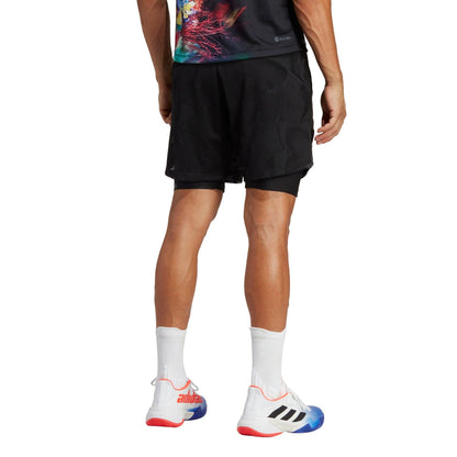 A person stands facing away, wearing a colorful T-shirt and adidas Melbourne 2in1 Men's 7 Inch Tennis Shorts in black, featuring AEROREADY technology. They are complemented by white athletic shoes with red and blue accents on the soles and black stripes on the sides, paired with white socks.