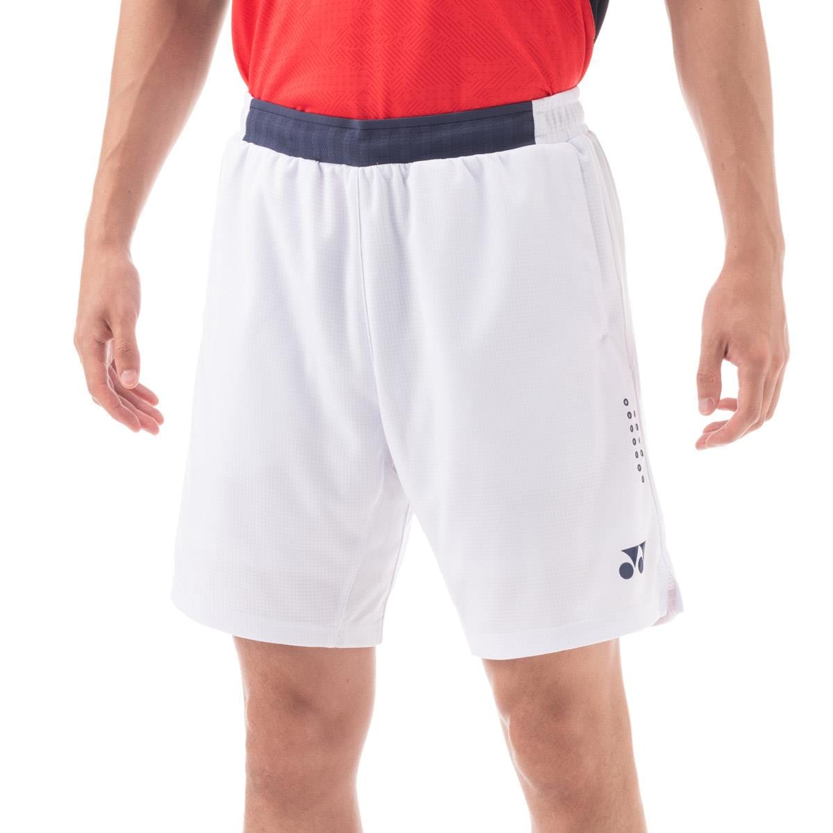 A person stands in a relaxed pose, showcasing Yonex 15131 Mens Shorts - White, designed with VERY COOL technology. The red shirt pairs well with the white performance shorts that feature a navy waistband and visible logos. The focus is on the lower half of the body, highlighting both style and innovation.