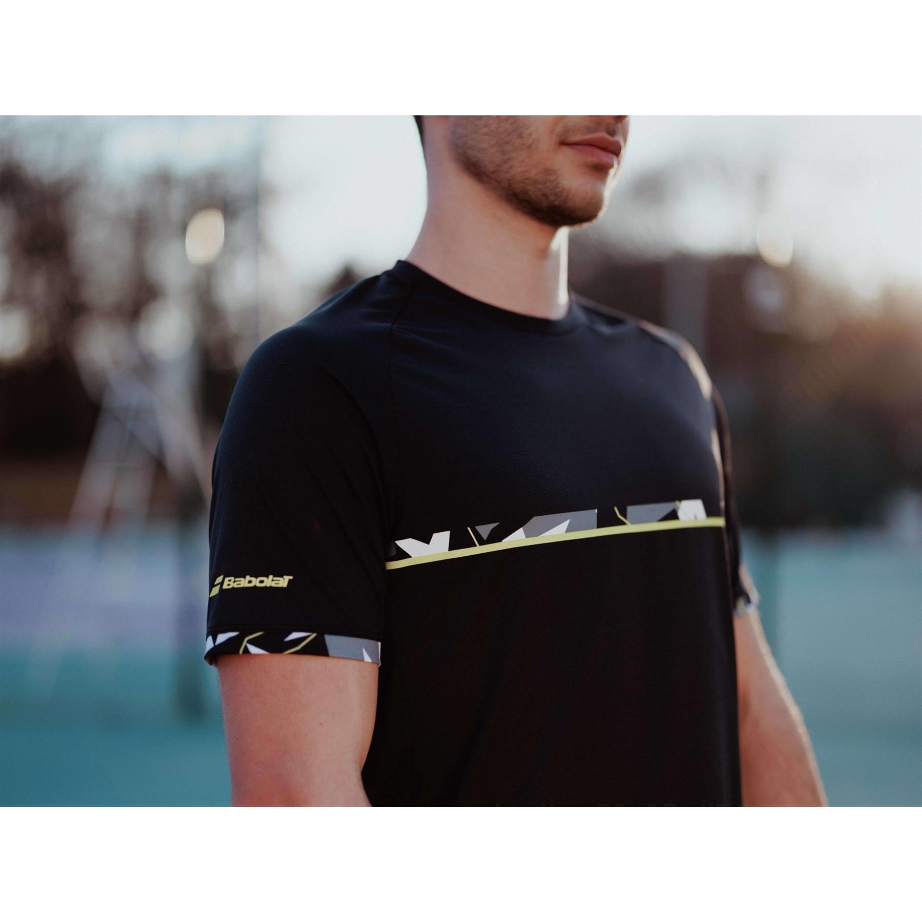 A person wearing the stylish Babolat Aero Crew Neck Men's Tennis T-Shirt in black, showcasing its breathable fabric with geometric patterns and the iconic "Babolat" logo on the sleeve, stands outdoors against a blurred backdrop of a tennis court and trees—ideal for enhancing performance in racket sports.