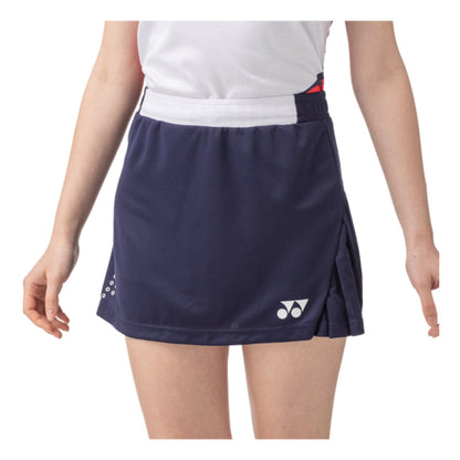 A person is wearing a navy blue Yonex 26094 Women's Skort that showcases the brand's logo near the bottom. The pleated design not only adds style but also provides UV protection. Paired with a white top, this skort incorporates VERY COOL TECHNOLOGY. The person's hands rest at their sides.