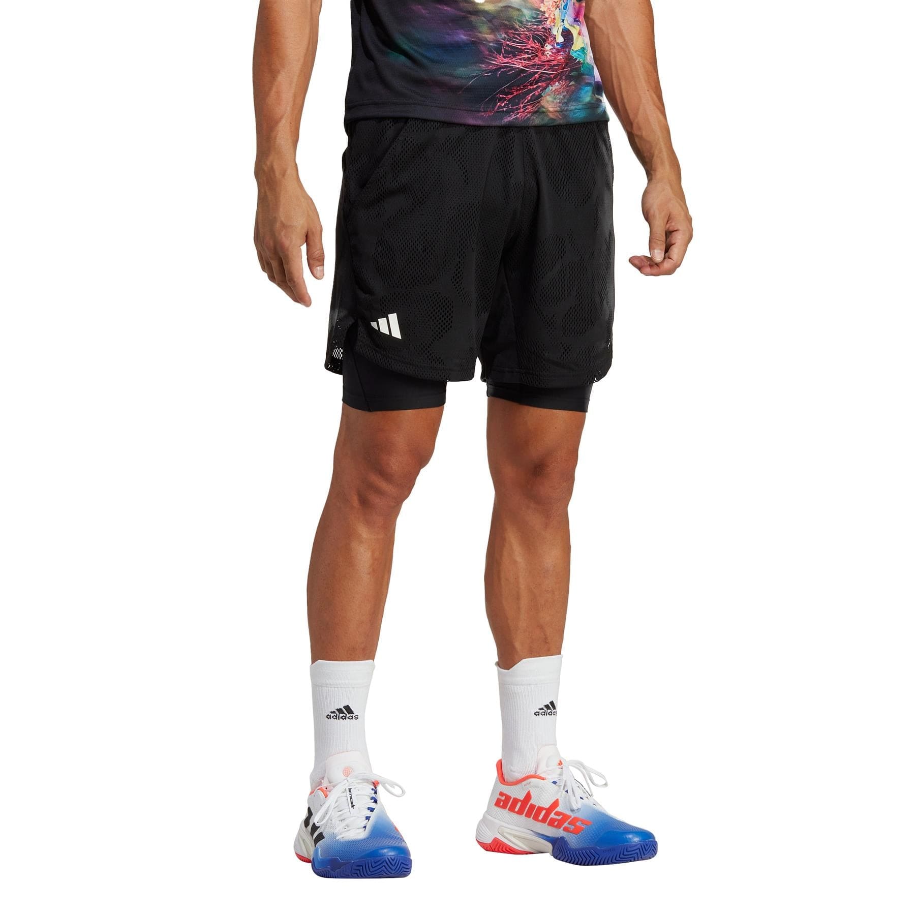 A man wearing a vibrant shirt and black adidas Melbourne 2in1 Men's 7 Inch Tennis Shorts stands with one visible hand, donning white AEROREADY socks and sneakers with blue and red accents. Only the lower half of his body is shown against a pristine white background.