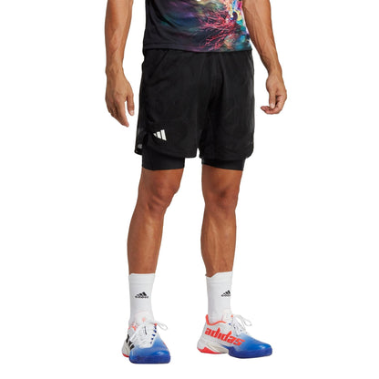 A man wearing a vibrant shirt and black adidas Melbourne 2in1 Men's 7 Inch Tennis Shorts stands with one visible hand, donning white AEROREADY socks and sneakers with blue and red accents. Only the lower half of his body is shown against a pristine white background.
