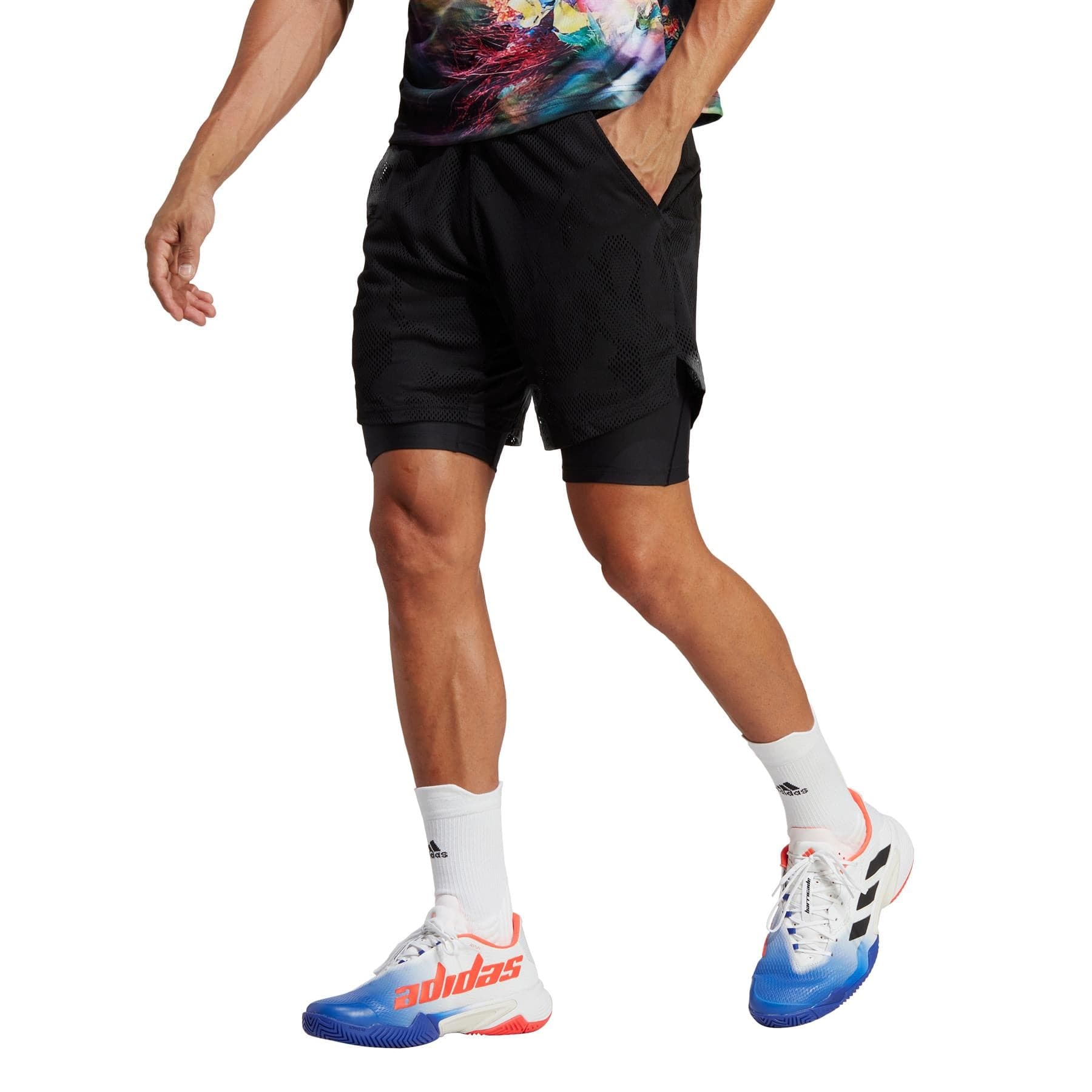 A person, wearing a colorful shirt and adidas Melbourne 2in1 Men's 7 Inch Tennis Shorts in black, along with mismatched red and blue sneakers, is walking. The lower body is highlighted against a crisp white background.